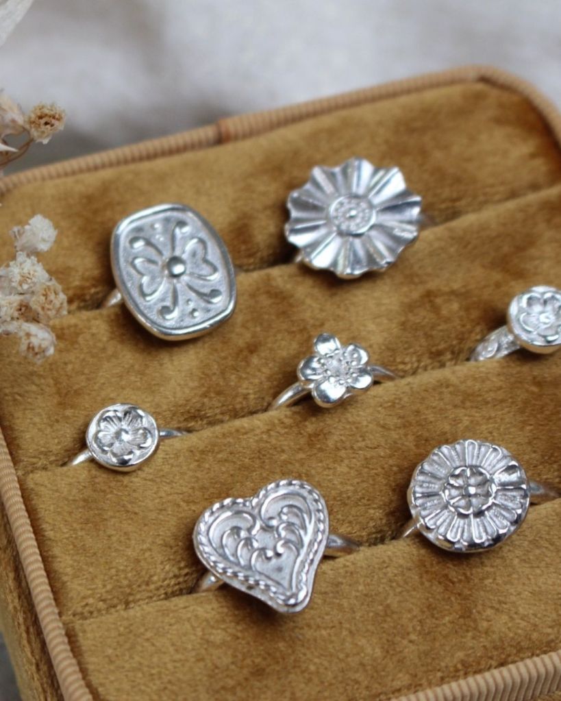 A collection of handmade silver rings displayed on a soft, brown velvet cushion, featuring floral and heart designs.