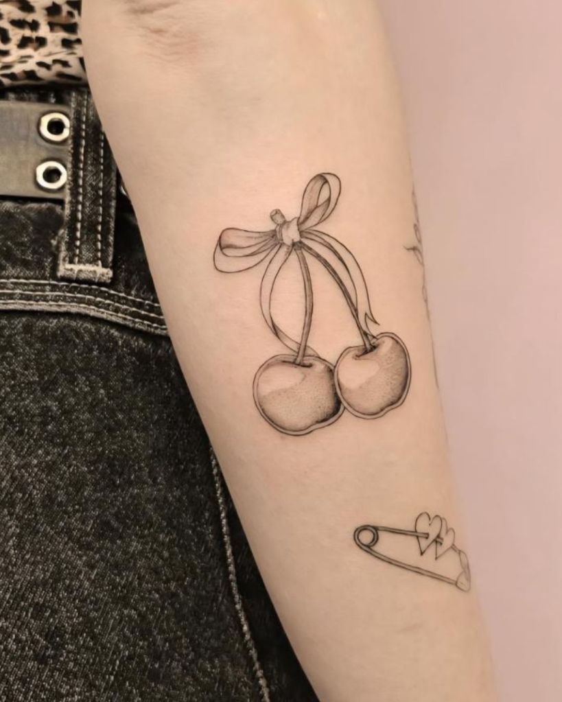 Close-up of a person's forearm showing two black outlined cherry tattoos tied with a ribbon and a small safety pin tattoo.